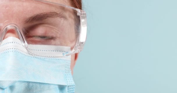 Close-up de um médico ou médico em equipamentos de proteção individual e óculos transparentes e luvas cirúrgicas azuis olhando para a câmera em um fundo azul — Vídeo de Stock
