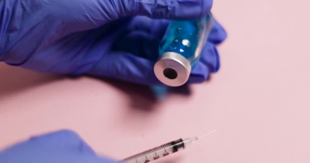 Kvinna hand i blå latexhandske håller sprutan injektion och glasflaska med flytande vaccin på rosa bakgrund — Stockvideo