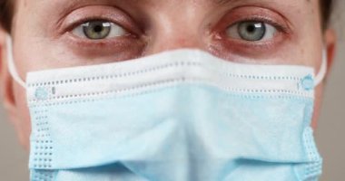 Yeşil gri gözlü kadın kameraya bakıyor ve göz kırpıyor. Koruyucu mavi maskeli kadın doktor gribe karşı korunuyor.
