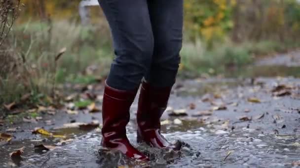 Gros plan d'une femme en bottes en caoutchouc rouge sautant dans une flaque boueuse d'automne après la pluie — Video