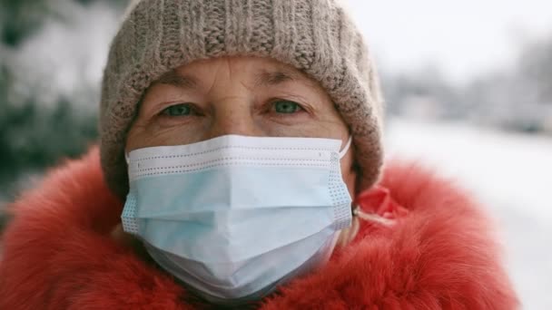 Volwassen oudere vrouw met een beschermend medisch masker kijkend naar de camera in het winterpark — Stockvideo