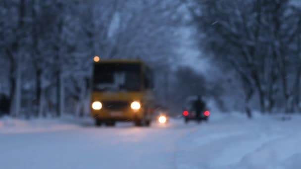 Voitures et route dans la neige après une chute de neige. Silhouette floue de bus jaune, voitures avec phares sur une route enneigée dans un parc au crépuscule — Video