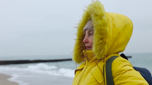 Donna anziana turista in una calda giacca gialla e cappello a maglia con uno zaino viaggia e cammina lungo la spiaggia vicino al mare. Tempesta forte e freddo. Viaggi e turismo. — Video Stock