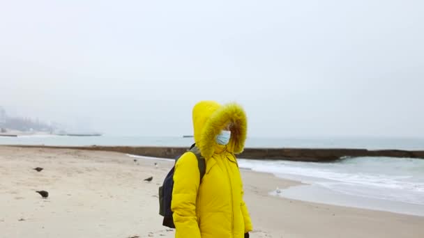 En vuxen kvinna i en varm gul jacka med ryggsäck använder en skyddande medicinsk mask, promenader längs stranden och njuta av en vacker utsikt över havet. Övergripande bild av pandemin COVID-19 — Stockvideo