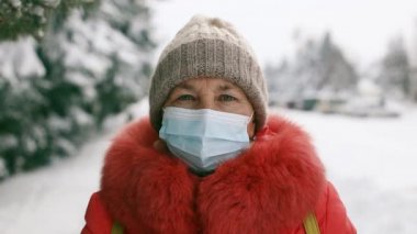 Kış parkındaki kameraya bakan koruyucu tıbbi maskeli yetişkin bir kadın. Coronavirus Covid 19 salgını sırasında sosyal mesafeler