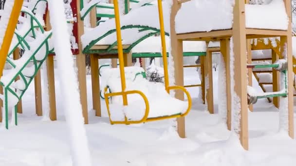 Lege kinderspeelplaats met glijbanen en schommels tijdens een sneeuwval in het park in de winter. Met sneeuw bedekte bomen. — Stockvideo