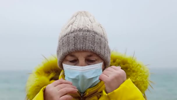 COVID-19 Pandemic coronavirus senior woman tourist in warm jacket with a backpack travel and walks along the beach near the sea. Fuerte tormenta y clima frío. Viajes y turismo. — Vídeo de stock