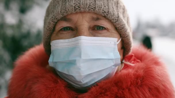 Donna anziana adulta in una maschera medica protettiva guardando la fotocamera nel parco invernale durante la pandemia di Coronavirus Covid 19 — Video Stock