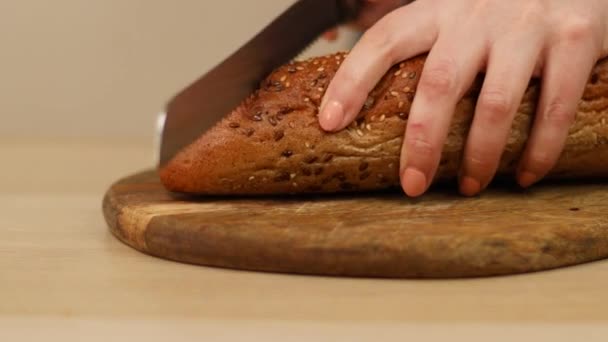 Skär en limpa surdegsbröd med vass kniv på ett träbord. Kvinnliga händer skär rågbröd med frön på träskiva. — Stockvideo