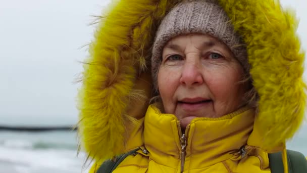 Sırt çantalı, sıcak, parlak elbiseli yaşlı bir turist kışın deniz kenarında yürür. Tatiller ve seyahat kavramı — Stok video