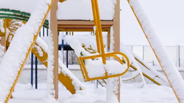 Пустые детские площадки с качелями во время снегопада. Зимой в парке заснеженные деревья — стоковое видео
