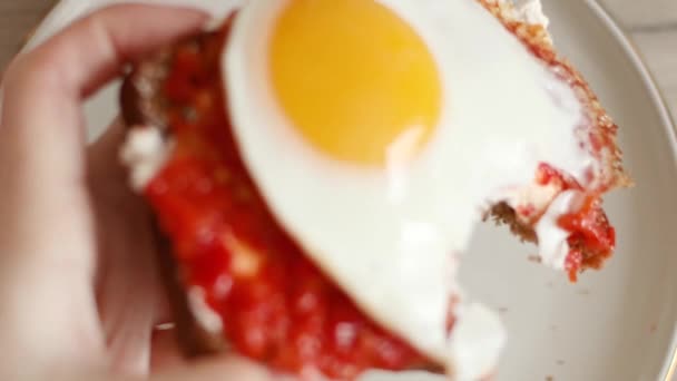 Kvinnliga händer biter av en bit smörgås med färskost, soltorkade tomater eller adjika och ett stekt ägg — Stockvideo