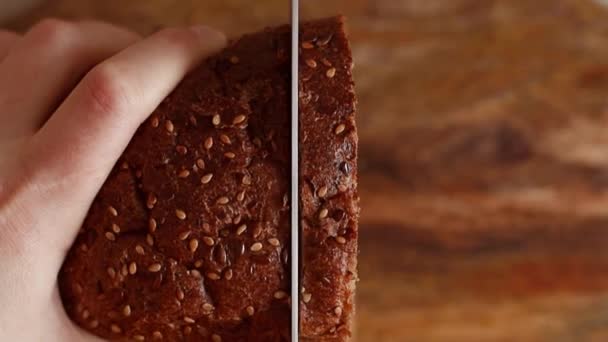 Mão feminina corta pão de centeio fresco com sementes em uma tábua de madeira na cozinha com uma faca de metal afiada — Vídeo de Stock