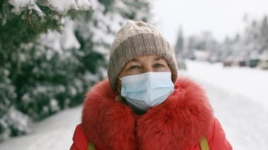 Kış parkındaki kameraya bakan koruyucu tıbbi maskeli yetişkin bir kadın. Coronavirus Covid 19 salgını sırasında sosyal mesafeler