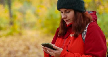 Sırt çantalı genç bir yürüyüşçü kız telefonuna güzel bir sonbahar ormanı patikasında mesaj yazıyor.