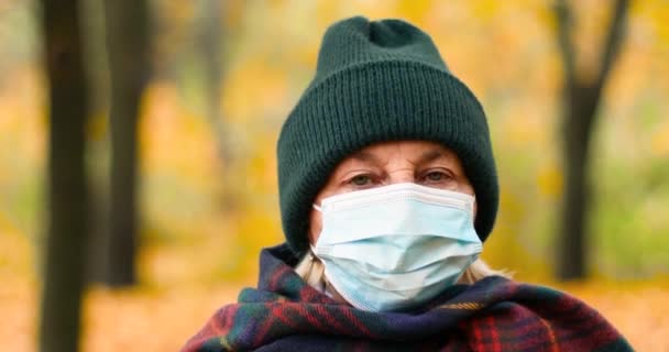 Seorang wanita tua berusia 50-an, mengenakan pakaian hangat dan topeng pelindung medis, melihat ke kamera. Berjalan di taman di hutan musim gugur. — Stok Video