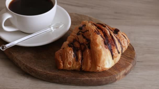 Americano koffie in keramische beker met verse gebakken chocolade croissant op houten plank. Het nemen van een croissant van houten plank. — Stockvideo