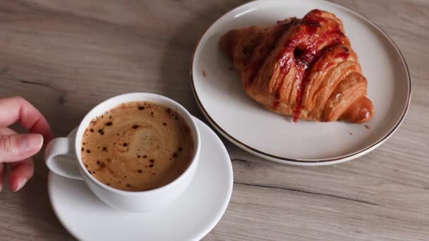 Lahodný čerstvý francouzský croissant s jahodovou náplní a šálkem aromatické kávy na dřevěném stole. Vzít si šálek kávy ze stolu ze dřeva. — Stock video