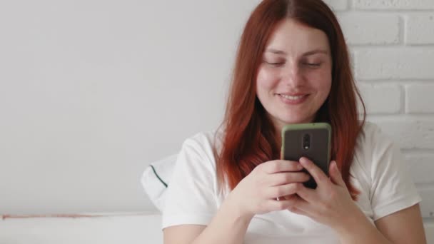 Позитивная молодая кавказская рыжая девушка, сидящая в постели и пользующаяся смартфоном. Доброе утро. — стоковое видео