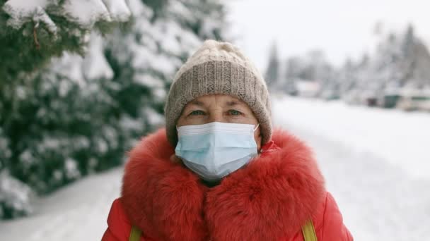 Eine erwachsene Seniorin in medizinischer Schutzmaske blickt im Winterpark in die Kamera. Soziale Distanzierung während der Coronavirus Covid 19 Pandemie — Stockvideo