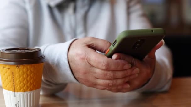 Женские руки используют смс-сообщения на мобильном телефоне в социальных сетях b, сидя в кафе. Бумажная чашка с кофе или горячим чаем на столе — стоковое видео