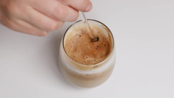 Fazendo café de manhã com o leite, mexe o açúcar em uma xícara. Mulher mão pessoa mexendo café latte com colher de vidro na mesa branca — Vídeo de Stock