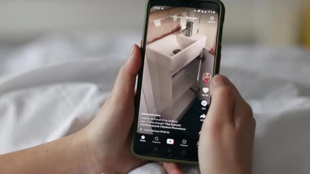 Девушка смотрит видео в популярном приложении Tiktok на черном смартфоне, лежащем на кровати дома — стоковое видео
