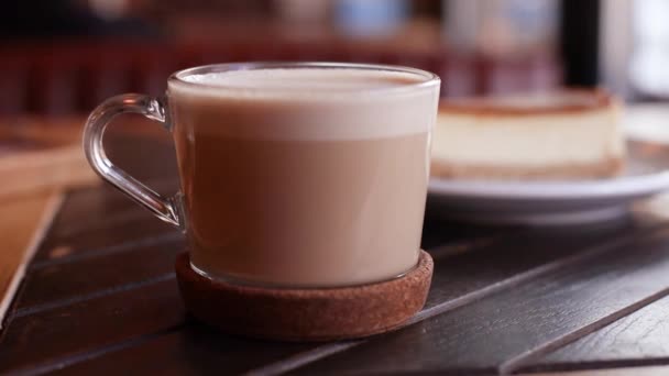 Primer plano de la taza de vidrio de café con leche caliente con un pastel de queso en el plato en la mesa — Vídeo de stock