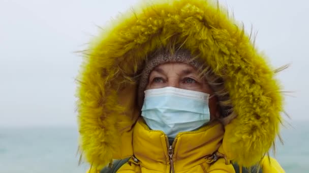 COVID-19 Pandemic coronavirus femeie turistică în vârstă într-o jachetă galbenă caldă și pălărie tricotată cu un rucsac călătorește și se plimbă de-a lungul plajei din apropierea mării. Furtună puternică şi vreme rece. Călătorii și — Videoclip de stoc