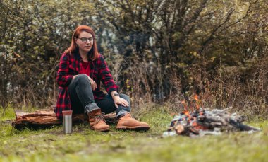 Dişi gezgin ormanda kamp yapıyor ve yorucu bir günün ardından ateşin başında dinleniyor. Termosta sıcak çay ya da kahve tutan bir kız. Seyahat, macera ve mevsimlik tatil konsepti.