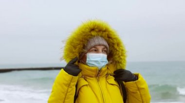 COVID-19 Pandemic Coronavirus kıdemli kadın turist sırt çantalı ve deniz kenarında yürüyor. Güçlü fırtına ve soğuk hava. Maske takan kişi