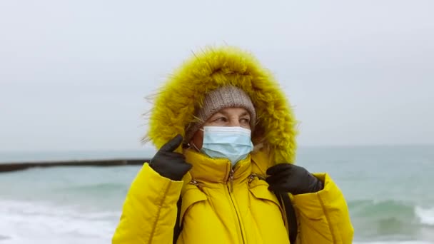 COVID-19 Pandemic coronavirus senior woman tourist in warm jacket with a backpack travels and walks along the beach near the sea. Strong storm and cold weather. Person wearing mask protective for — Stock Video