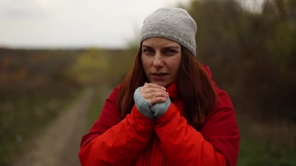 El primer plano de una niña con una chaqueta deportiva roja y un sombrero ruboriza las manos congeladas en el bosque de otoño. Concepto de camping, turismo de naturaleza. — Vídeos de Stock