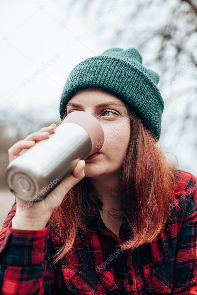 Caucasian woman drinks from a mug of thermos. Rest at in nature autumn forest