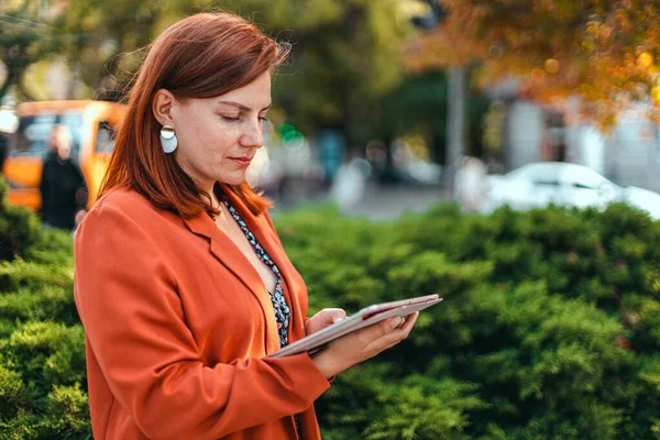 Mulher de negócios profissional bonita em roupas de escritório elegantes em suas mãos usa um dispositivo tablet em um parque da cidade na cidade — Fotografia de Stock