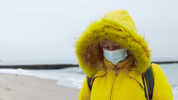 Mujer adulta en una chaqueta de color amarillo cálido con una mochila utiliza una máscara médica protectora, paseos a lo largo de la orilla del mar y disfrutar de una hermosa vista del mar. Imagen global de la pandemia de COVID-19 — Vídeos de Stock