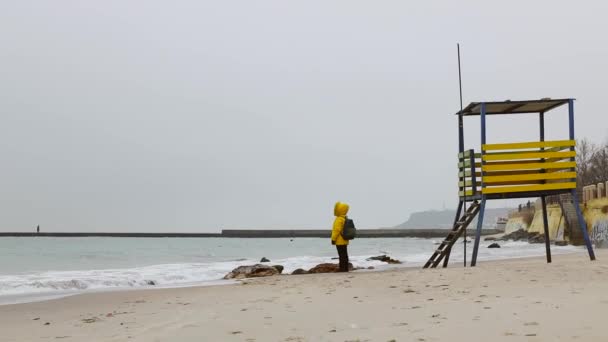 Un hombre con una chaqueta de color amarillo cálido con una mochila se para en la orilla del mar y mira a la distancia disfrutando de la soledad. Viajes y turismo. — Vídeos de Stock