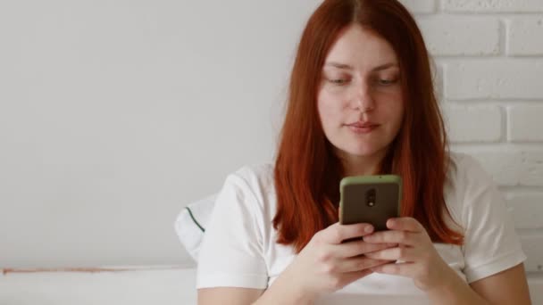 Uma jovem caucasiana positiva de cabelos ruivos usando telefone em casa na manhã. Bom dia. — Vídeo de Stock