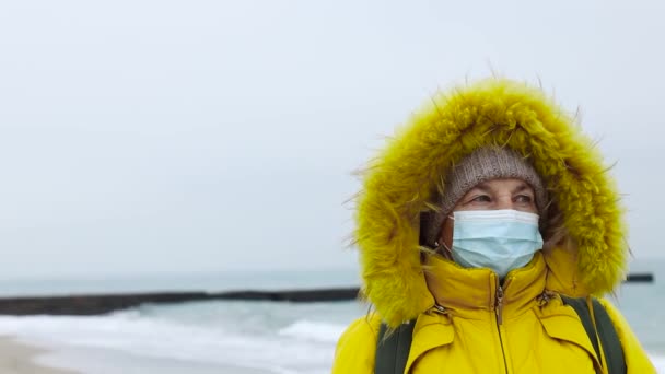 An adult woman in a warm yellow jacket with a backpack in a medical protective mask walks along the seashore and looks into the distance enjoying the beautiful sea view — Stock Video