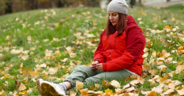 Fiatal, gyönyörű kaukázusi lány meleg ruhában és kalapban ül a fűben, lehullott levelekkel a parkban. Lány kéz kezében egy lehullott levél — Stock videók