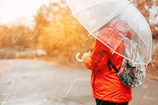 Una mujer con una mochila y una chaqueta roja camina por las calles húmedas y lluviosas bajo un paraguas transparente. Vista posterior con espacio de copia — Foto de Stock