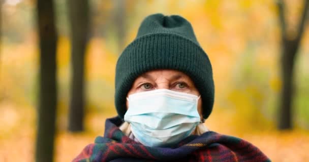 An elderly woman 50s, wearing warm clothes and a protective medical mask, looks at the camera. Walking in a park in an autumn forest. — Stock Video