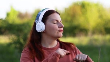 Neşeli, genç, beyaz bir kadın kulaklıkla müzik dinliyor, dans ediyor, şarkı söylüyor, eğleniyor, elleriyle el kol hareketi yapıyor.