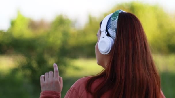 Achteraanzicht van een blanke vrolijke vrouw met een witte hoofdtelefoon die naar muziek op straat luistert en naar muziek buiten luistert. — Stockvideo