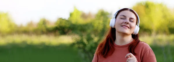 Hermosa joven con los ojos cerrados escuchando música a través de auriculares y bailando en el día soleado en el parque de la ciudad — Foto de Stock