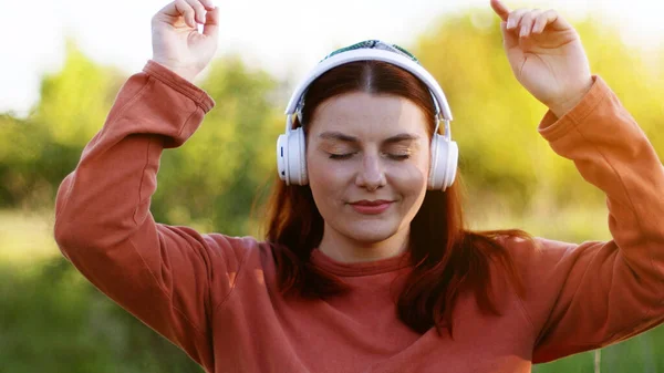 Vacker ung flicka med slutna ögon lyssna musik genom hörlurar och dansa på solig dag i stadsparken — Stockfoto