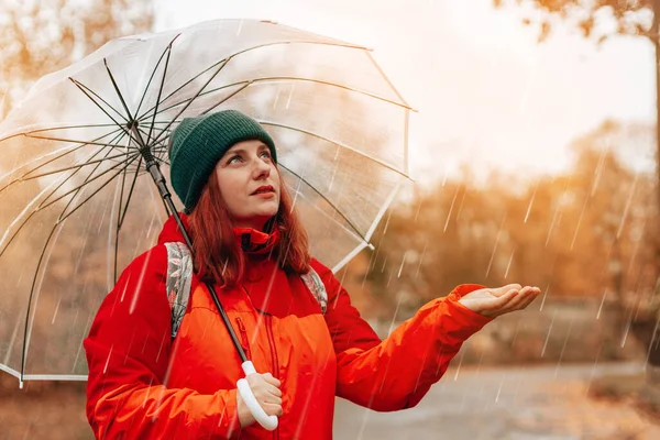 Portrait of caucasian woman with backpack and in red jacket walks along wet rainy streets under a transparent umbrella — Stock Fotó