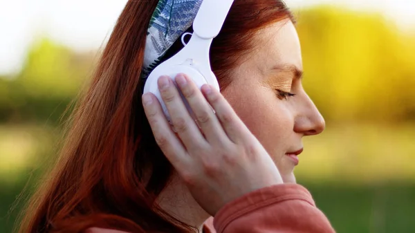 Attractive smart young woman listening music with headphones on sunny day while walking in city park — Stockfoto