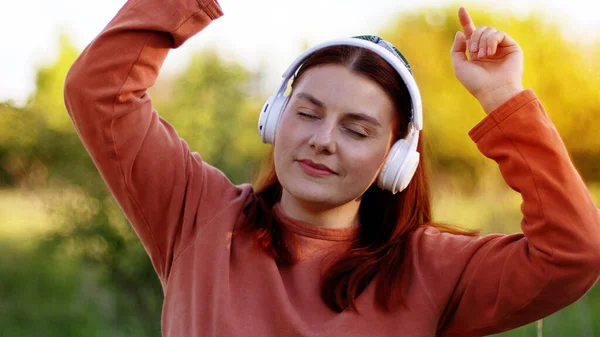 Vacker ung flicka med slutna ögon lyssna musik genom hörlurar och dansa på solig dag i stadsparken — Stockfoto