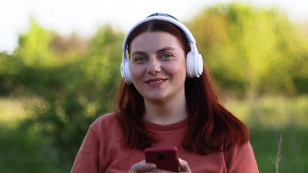 Beyaz çağdan kalma mutlu, gülümseyen bir kadın yüzü. Akıllı telefon kullanıyor ve kulaklıkla dışarıda sokakta müzik dinliyor. — Stok video